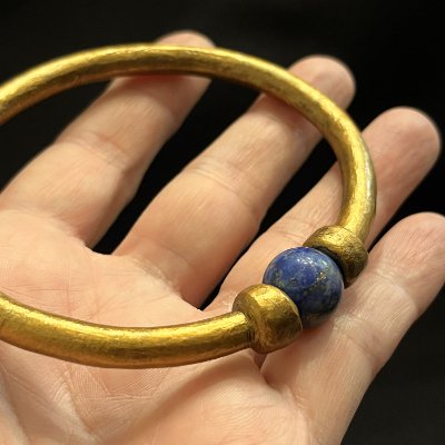 Detailed view of the gilded acrylic surface and lapis lazuli bead resting in an adult hand.