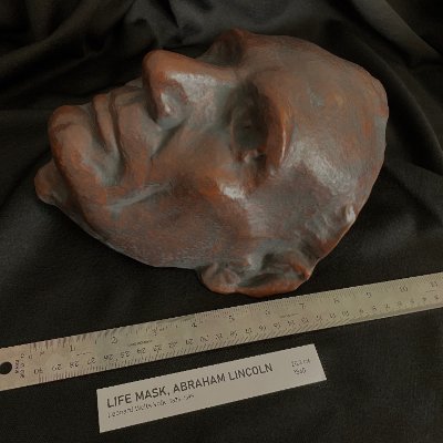 Right profile of the life mask displayed alongside a wooden ruler for size reference.