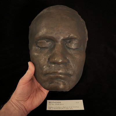 A hand supports the replica mask vertically to reveal its precise frontal symmetry.