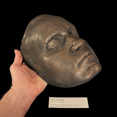 A hand holds the replica life mask at an angle to demonstrate its physical scale.