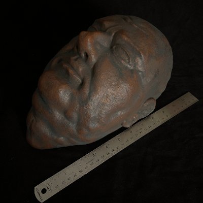 The death mask is positioned beside a metal ruler to document its physical length.