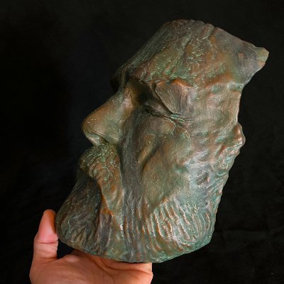 Angled frontal view of the patinated face mask held by a human hand to indicate scale.