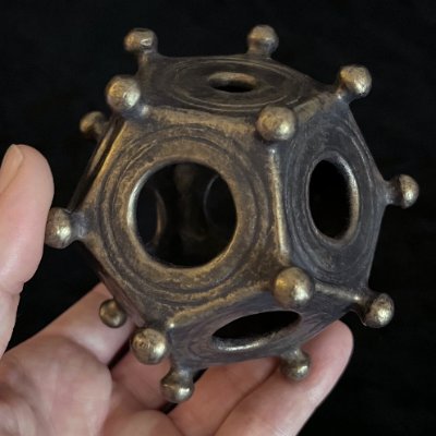 A hand holds the bronze-patinated Roman dodecahedron replica to demonstrate its physical scale.