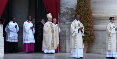 Pope Francis stands with attendants while holding the papal ferula outdoors.